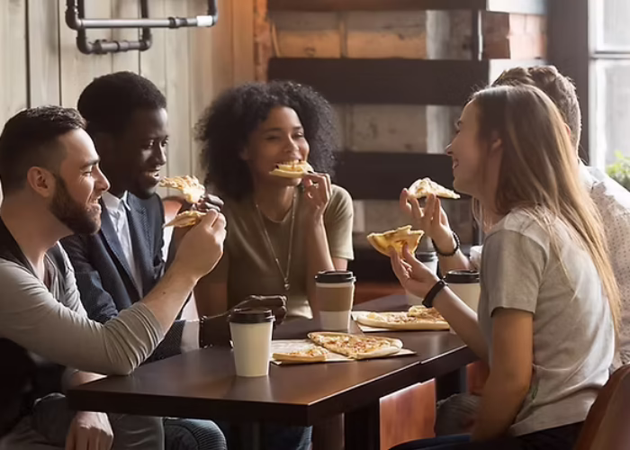 a group of friends smiling while eating pizza together in a restaurant